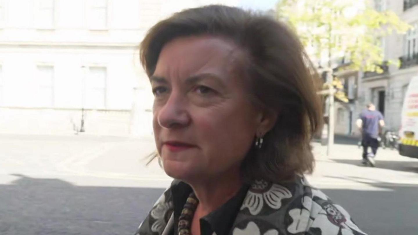 Eluned Morgan arriving at Lancaster House, wearing a grey and white top with flower patterns and a necklace. A man is out of focus in the background, walking away from the camera.