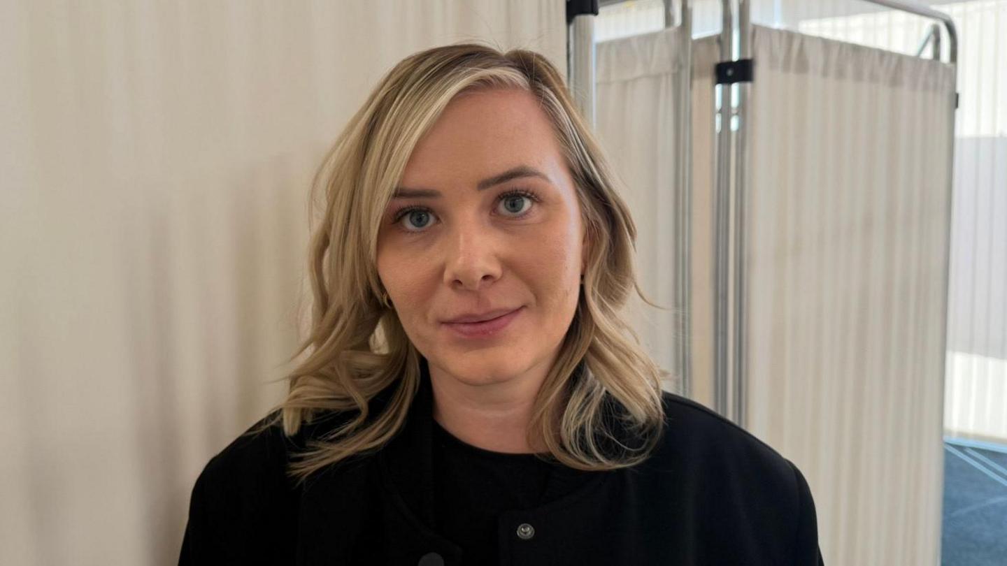 The picture shows a woman smiling at the camera. She has a short blonde bob, which is curled. She is also wearing a black jacket and is standing in front of a medical screen. 