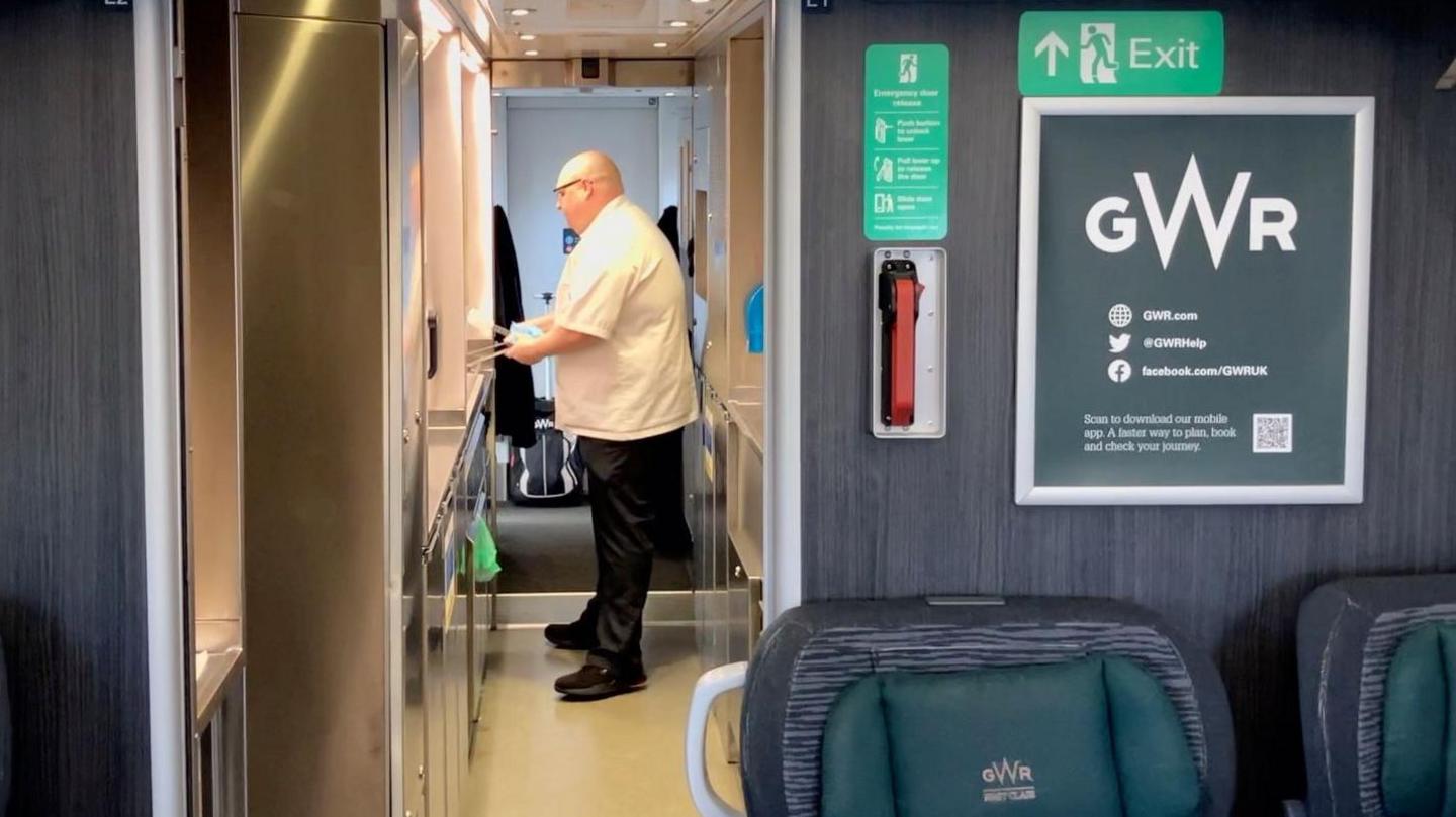 On board a train, a chef is standing in a corridor cooking pancakes. He wears a white chef's coat and black trousers and is bald and wears black glasses. He stands in a train kitchen.