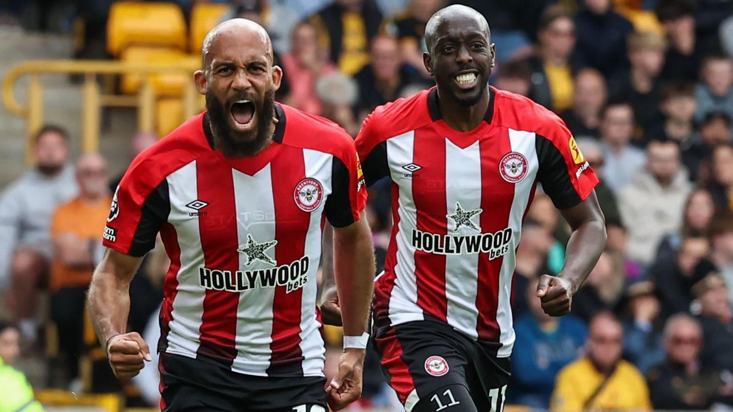 Bryan Mbeumo and Yoane Wissa seen running side by side after a goal for Brentford Football Club. Visible from their black shorts up, they are wearing the Bees' home jersey with wide red and white vertical stripes. The bearded Mbeumo, who is on the left and slightly nearer the camera, is opening his mouth to shout and clenching his right fist. Wissa on the right has his teeth clamped together in slightly rictus-like smile