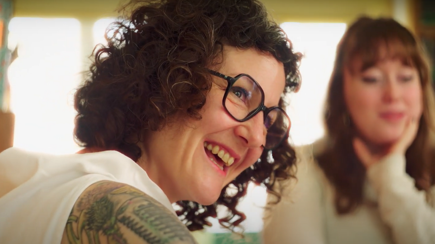 Imogen, who has dark curly hair and is wearing glasses, smiles. In the background a young woman can be seen resting her chin on her palm.