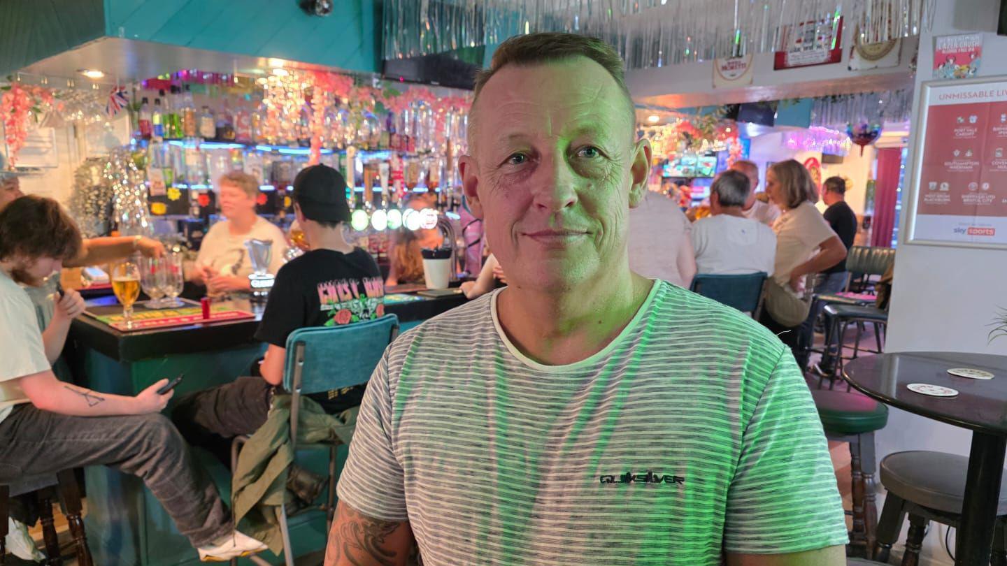 A man is pictured sat in a pub. He has brown hair and is wearing a white shirt with a black logo. Behind him people are sat around a bar.