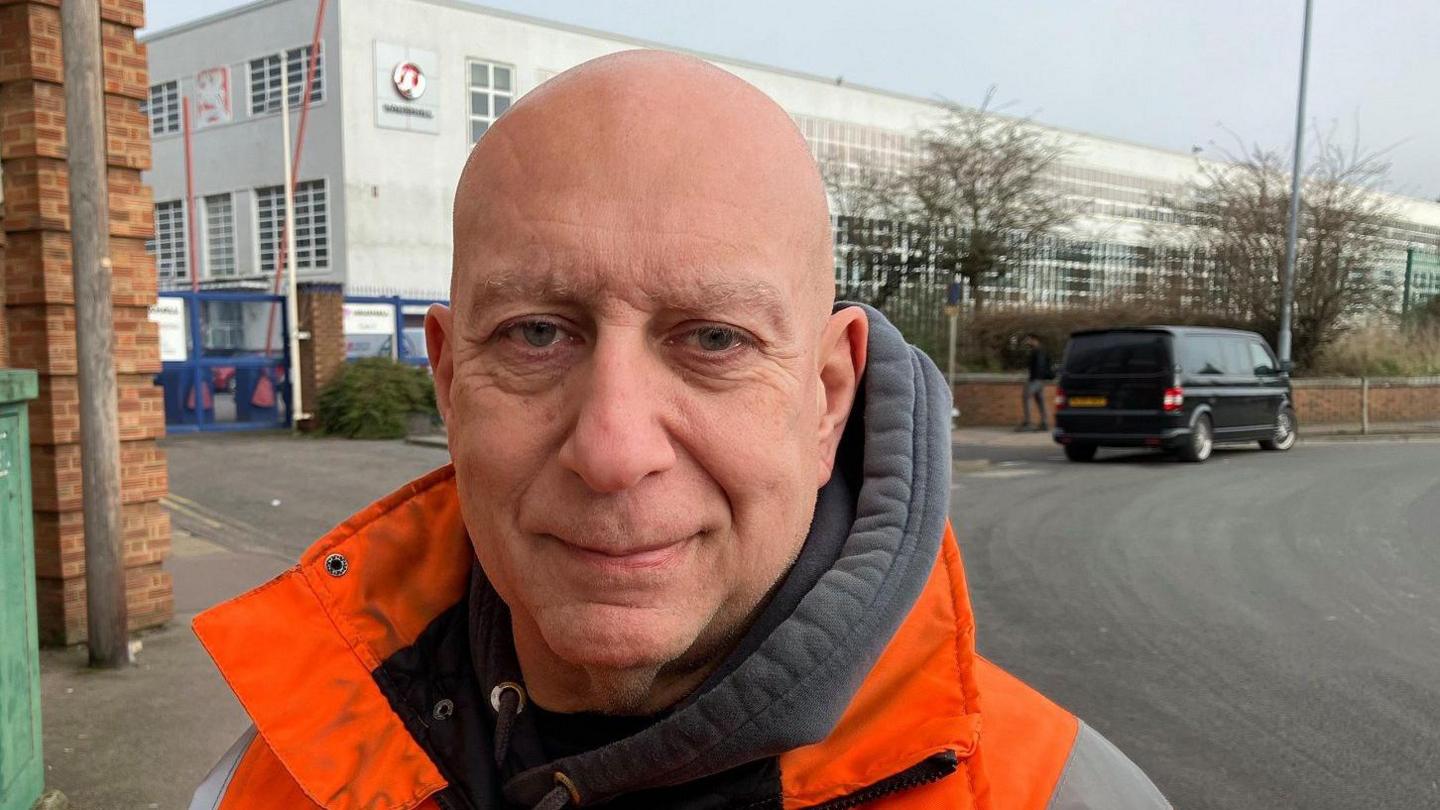 Dave wears a bright orange and grey work anorak and a grey hoodie. He is bald and stands next to the road outside the Vauxhall factory. The Vauxhall emblem is on one corner, and the building recedes into the distance with many windows across its two stories. 