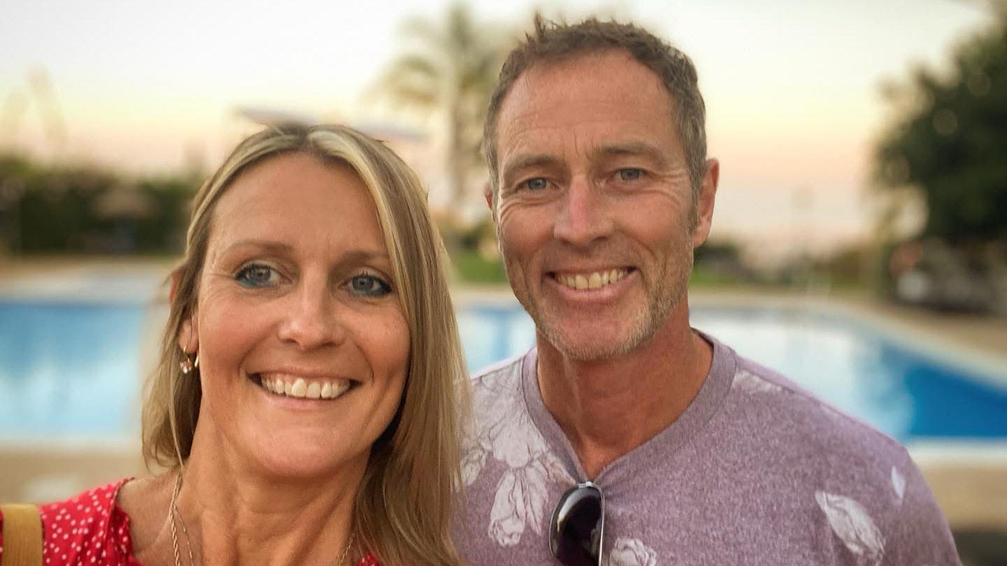 Lindsay Foreman and Craig Foreman smile as they pose together for a selfie picture. Behind them is a swimming pool. She is wearing a red top and him a lilac T-shirt.