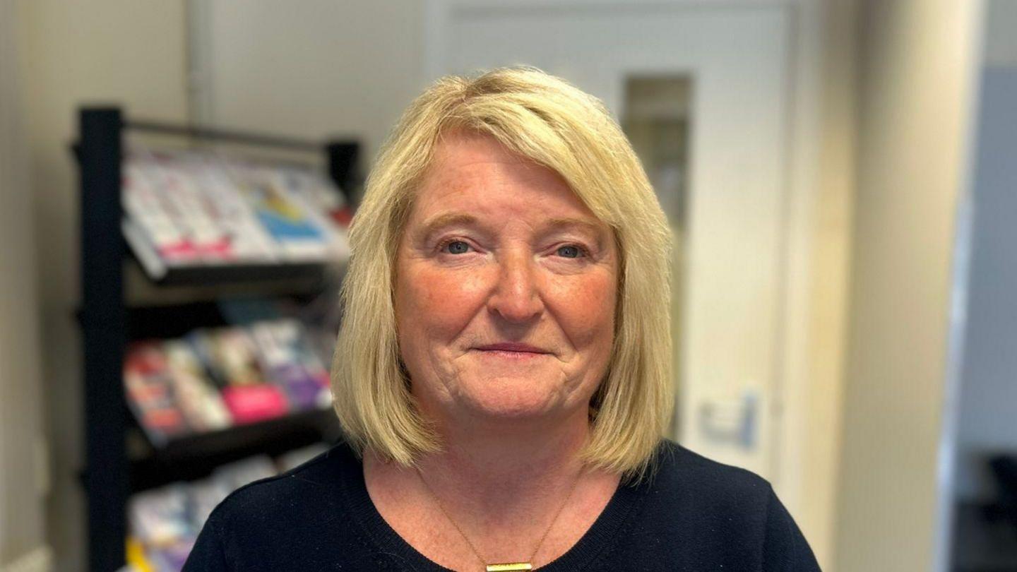 A woman with shoulder length blonde hair is looking into the camera. She's wearing a black round-neck top with a gold necklace.