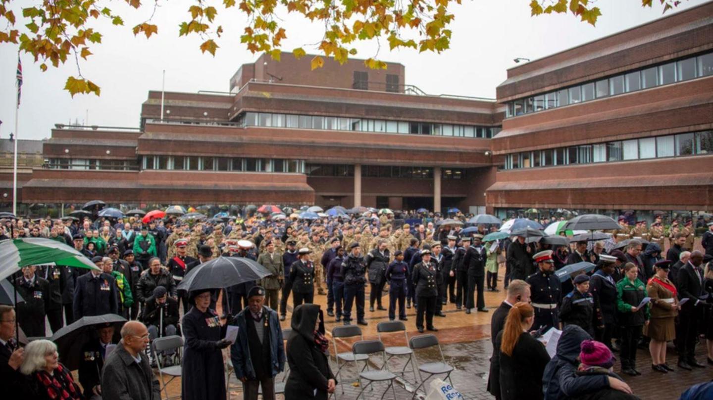 A large number of people outside council officers taking part in a remembrance event. Some people are holding umbrellas. There are members of armed forces in uniform alongside members of the public. 