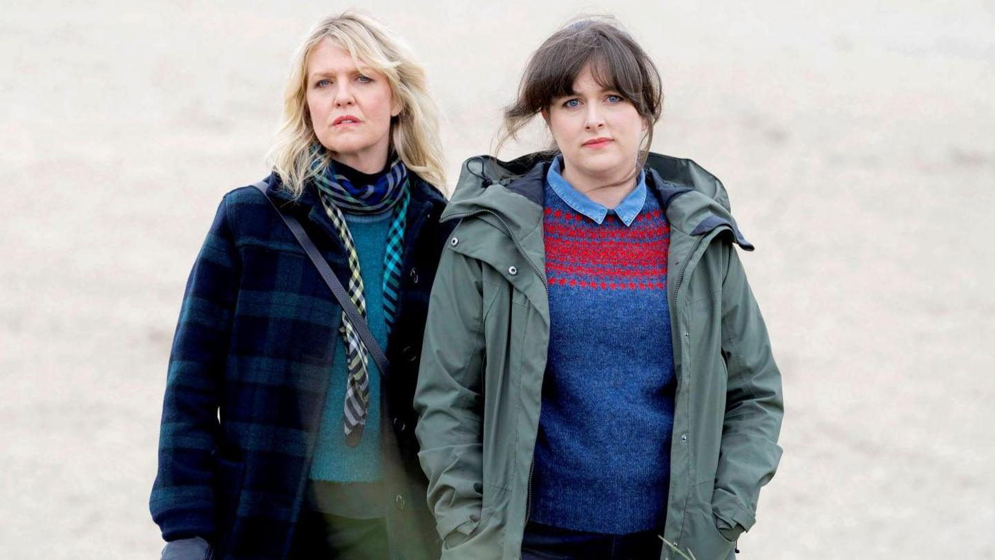 With the sandy beach stretching out behind them, a serious-looking DI Ruth Calder, in her dark coat and patterned scarf stands next to DI Tosh McIntosh, in a Shetland-patterned jumper and a waterproof khaki jacket with a hood