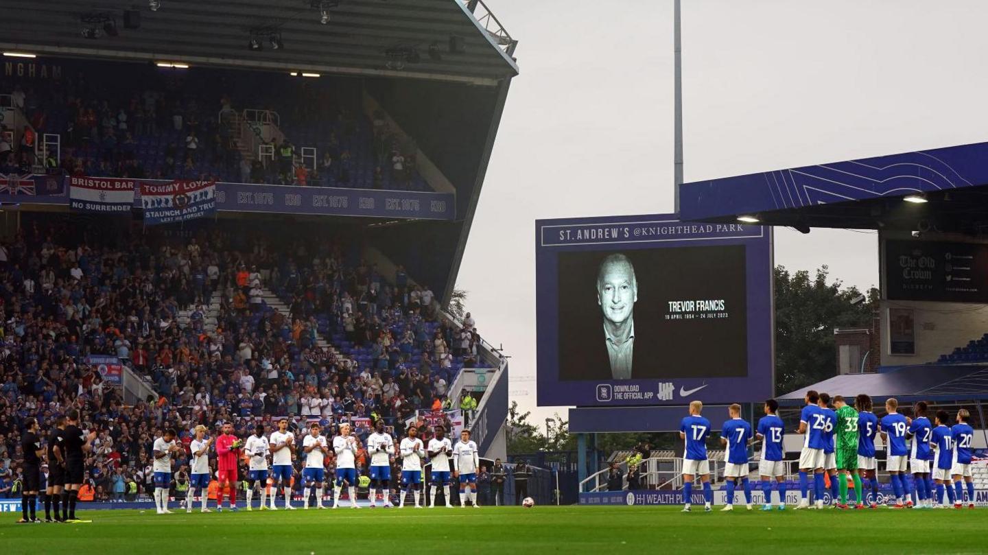 Two football teams stand on a pitch, with fans on seats in the background. The team on the left is wearing white, the team on the left is wearing blue. There is a large TV screen between the two teams with a black and white photo of a man and the words "Trevor Francis". 