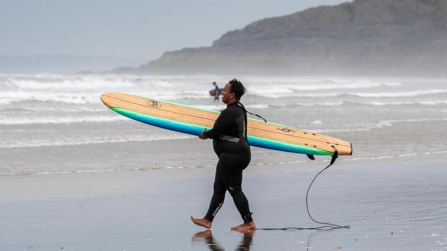 Jess Blake holding a foam board on her hip, she has the leash attached to her ankle and is wearing a wetsuit. In the background is the sea with a lot of white water waves and a headland jutting out into the sea. 