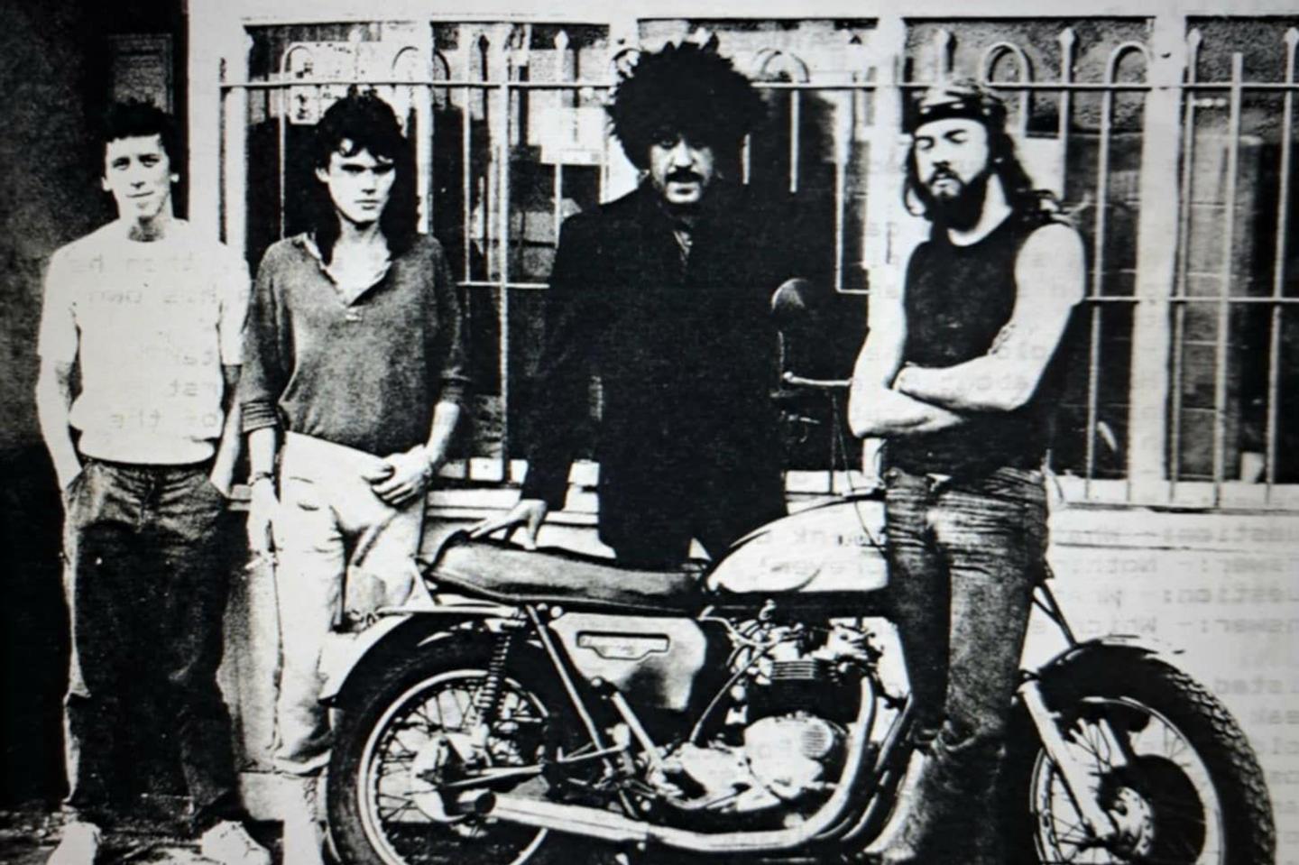 A moustachioed Phil Lynott with his distinctive shot of hair, and wearing a dark jacket, stands behind a motorcycle with Robin George to his right. George is wearing a jumper, and jeans. They are flanked by two other unnamed musicians. 