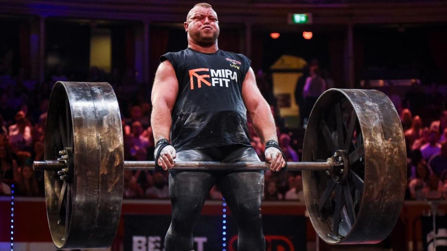 Kane Francis deadlifting during a competition. He is holding a metal barbell with huge metal plates that resemble steam roller wheels. His face is scrunched up and red from the exertion.