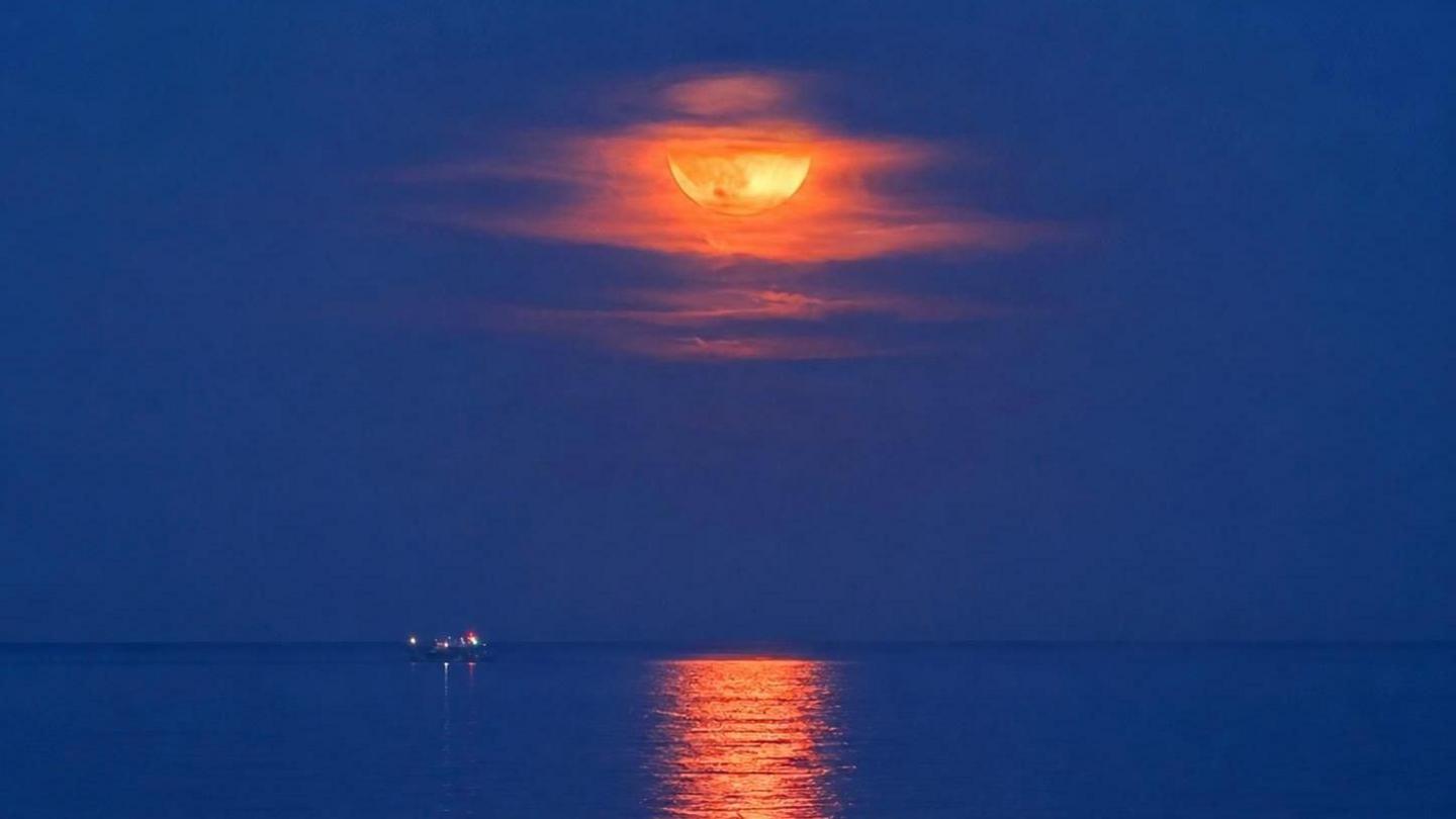 An orange hued full Strawberry Moon appears in a blue night sky above a still blue sea with a boat floating in it.  The sea reflects the moonlight.
