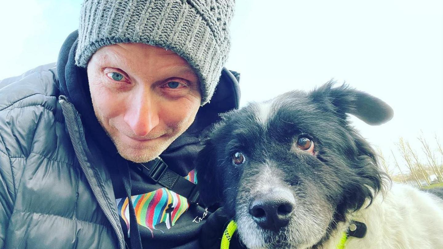 Dan Kay, wearing a grey woollen hat and grey puffer jacket, is photographed with his dog. 