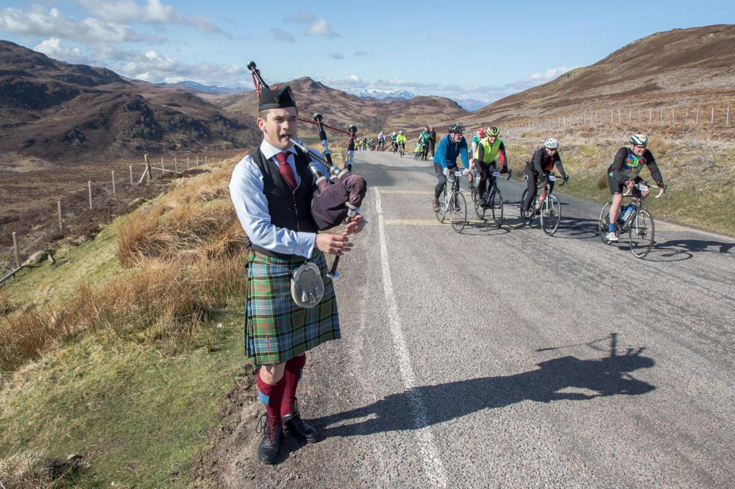 A piper wearing a kilt and Tam o' Shanter hat plays the bagpipes as cyclists ride past on a mountain road.