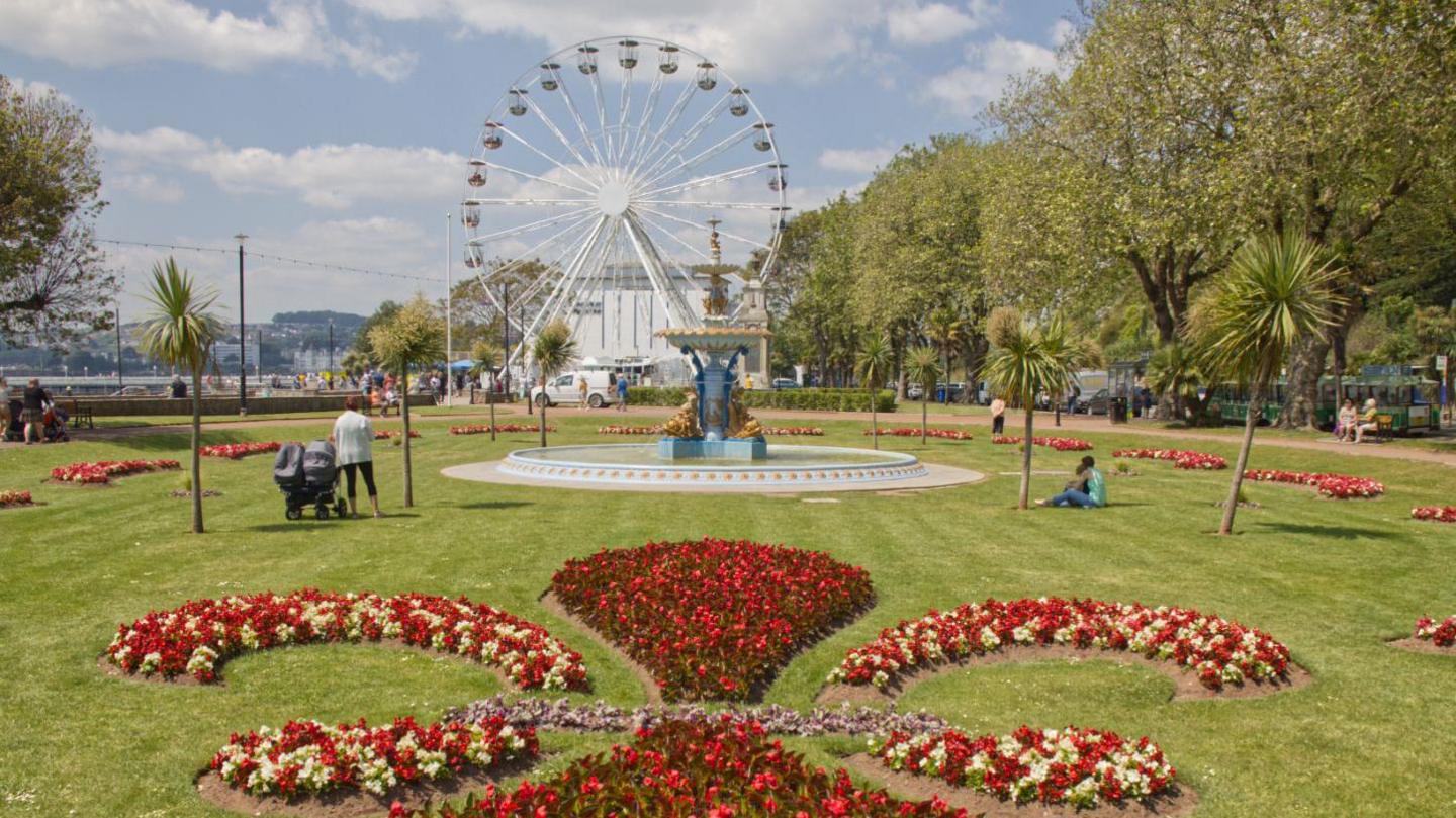 Princess Gardens and Royal Terrace Gardens, in the distance is a big wheel and in the foreground are a red flowers which have been arranged in an ornate style. There is a fountain in the middle of the image. The gardens are grassy and surrounded by trees on one side.