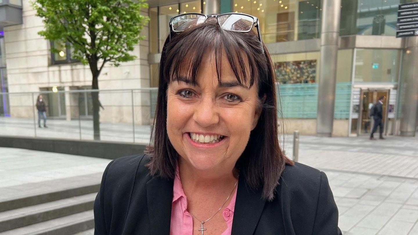 Lee-Bernadette Walford, a woman with brown hair with glasses on her head, is wearing a black suit jacket and pink blouse. She is stood outside a court building in Manchester.