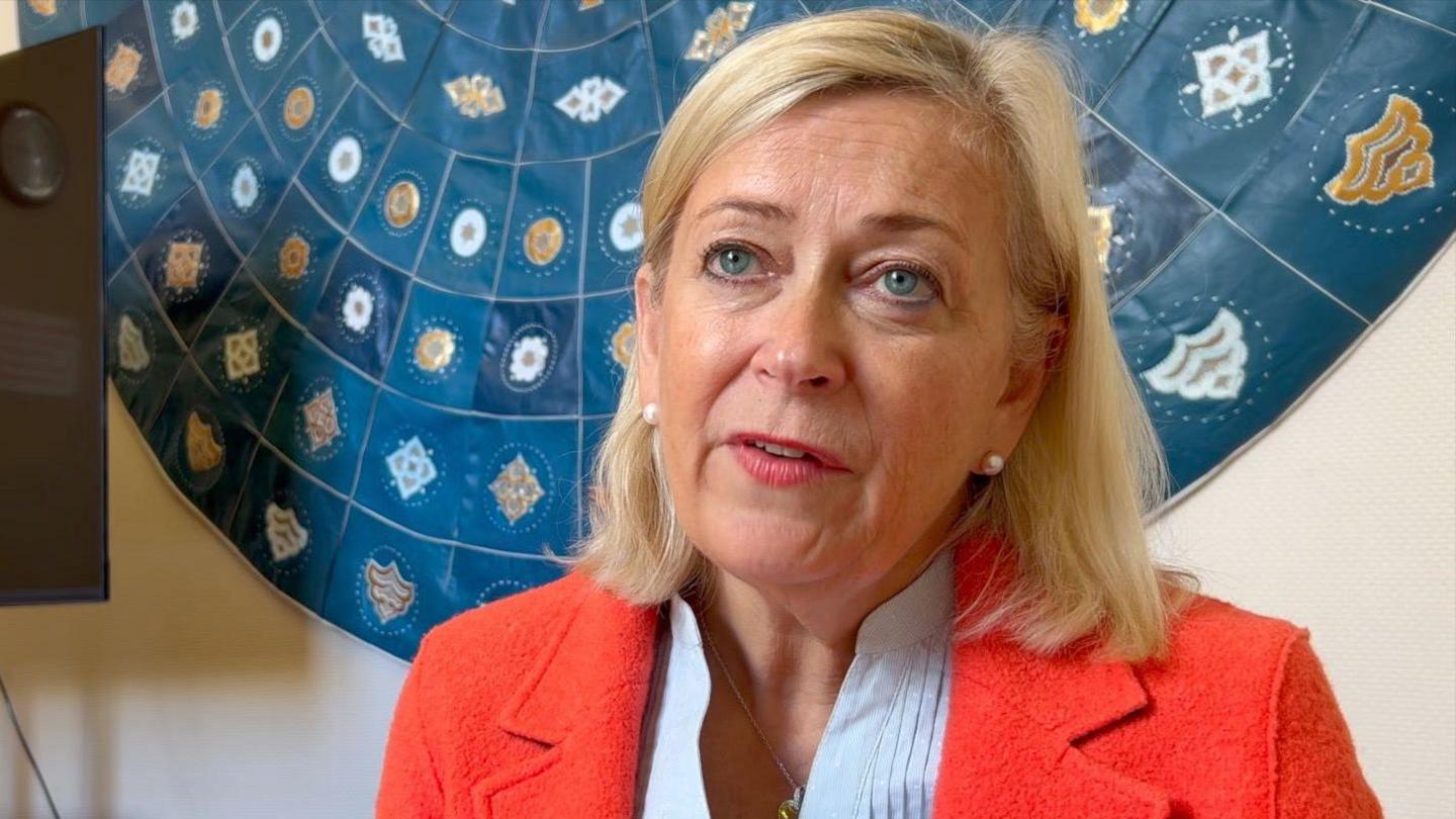 Helen has shoulder length blonde hair and is wearing a white shirt with a red blazer.