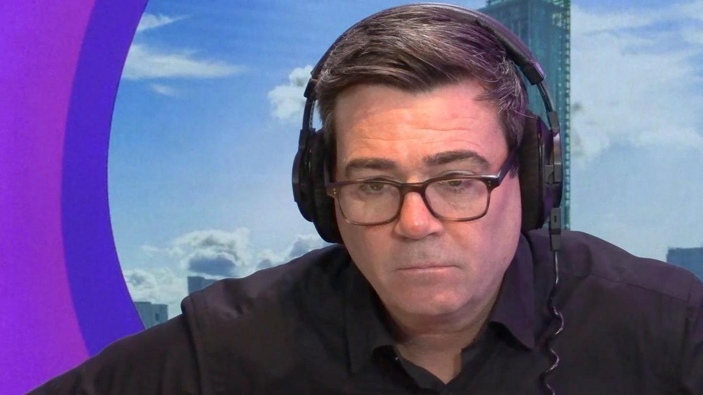 Andy Burnham wearing a black shirt and headphones, wearing glasses, in a radio studio. He is looking pensively towards the camera. 