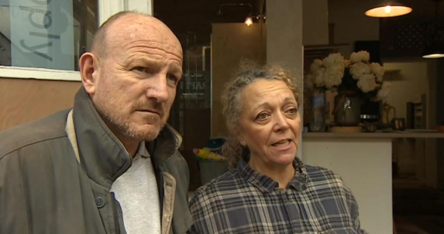 A man and woman at the entrance to a kitchen design shop. The man has a light beard, freckled face and is wearing a khaki coat and grey T-shirt. The woman has curly light brown hair tied back in a low ponytail and is wearing a blue and grey checked shirt. Flowers can be seen in a vase on a kitchen worktop in the background.