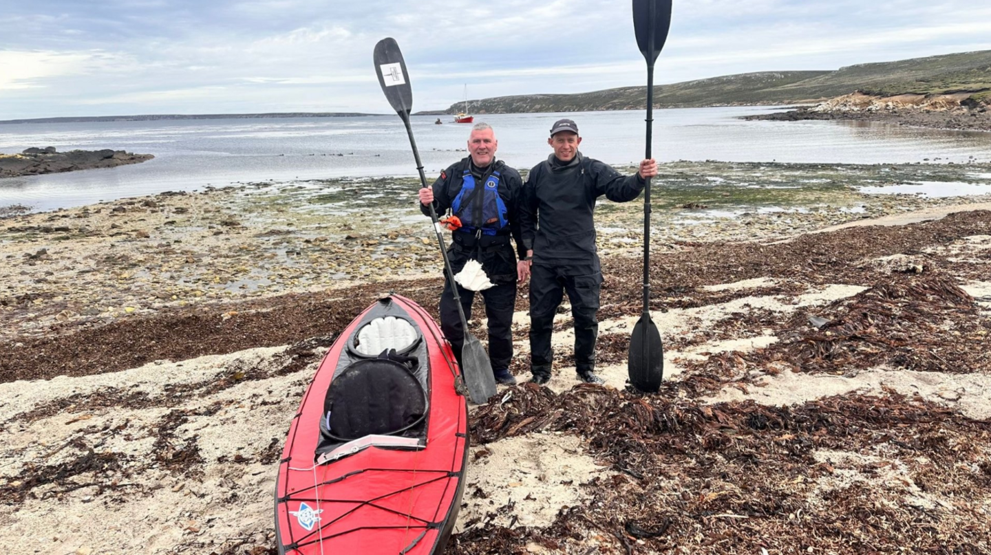 Image shows two men on the shore of a beach standing next to a red, two man kayak. They are holding their oars up next to them. 