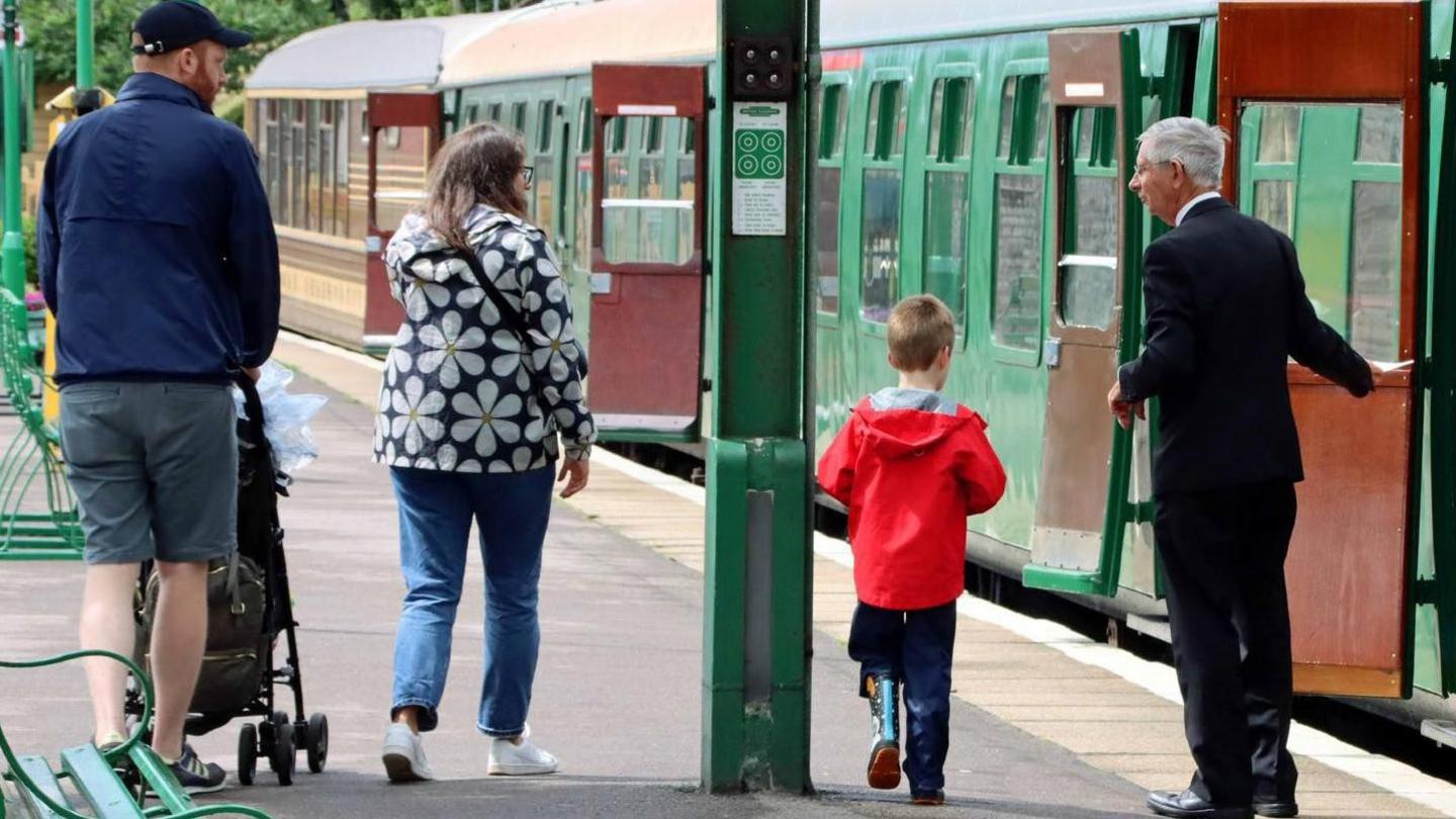 A family walk down a train platform next to a vintage train. A man is pushing a pushchair, and a woman and young boy walk alongside him. A train door is being held open by a man in a suit