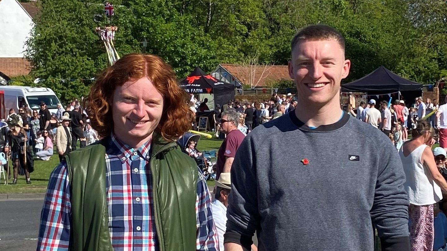 George Gilbert is wearing a navy shirt with large white and red checks and a dark green bodywarmer. Charlie is wearing a dark grey jumper. Behind the brothers - who are both smiling - are lots of people gathered on the village green during an event.