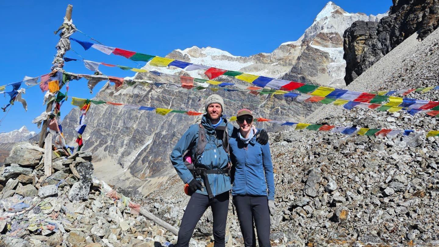 Rik stands next to Olivia on a mountain pass with his right arm around her shoulders. They are wearing blue jackets with black trousers. Rik has his beanie and Olivia is wearing a cap and sunglasses. There are colourful flags flying above their heads and in the background is Mount Everest.