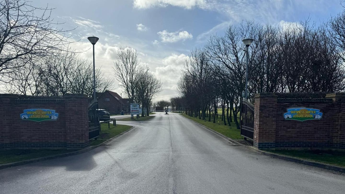 The entrance to Skegness Water Leisure Park has a tree-lined driveway. There are two black metal gates, which are open. There are brown brick walls with the leisure park logo, on both sides. The logo is blue, orange and green.