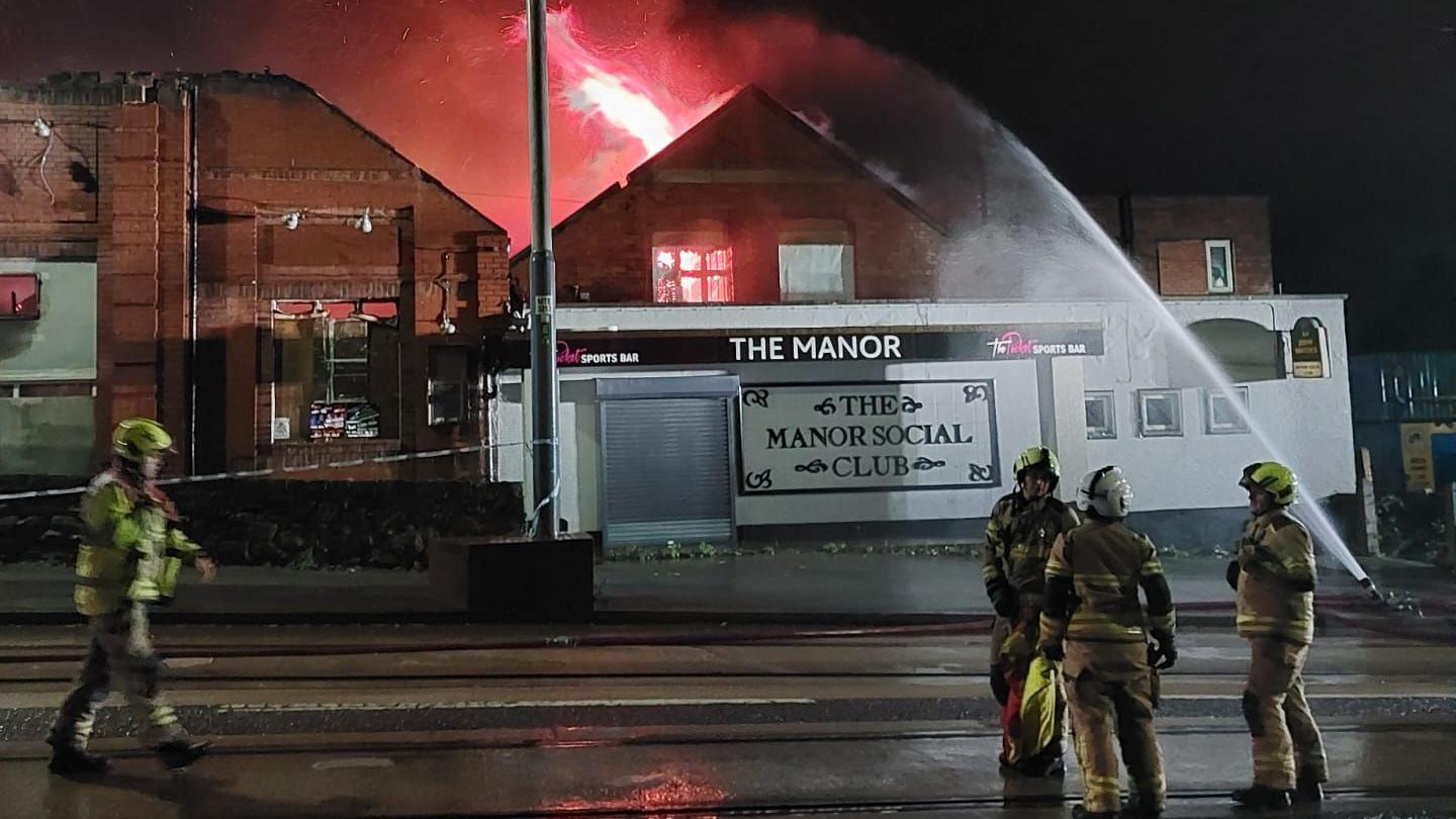 Four firefighters on the road outside a building which has a sign saying Manor Social Club on the front. A cordon can be seen tied to one side of the building. A jet of water is aimed at a large red flame within the building.