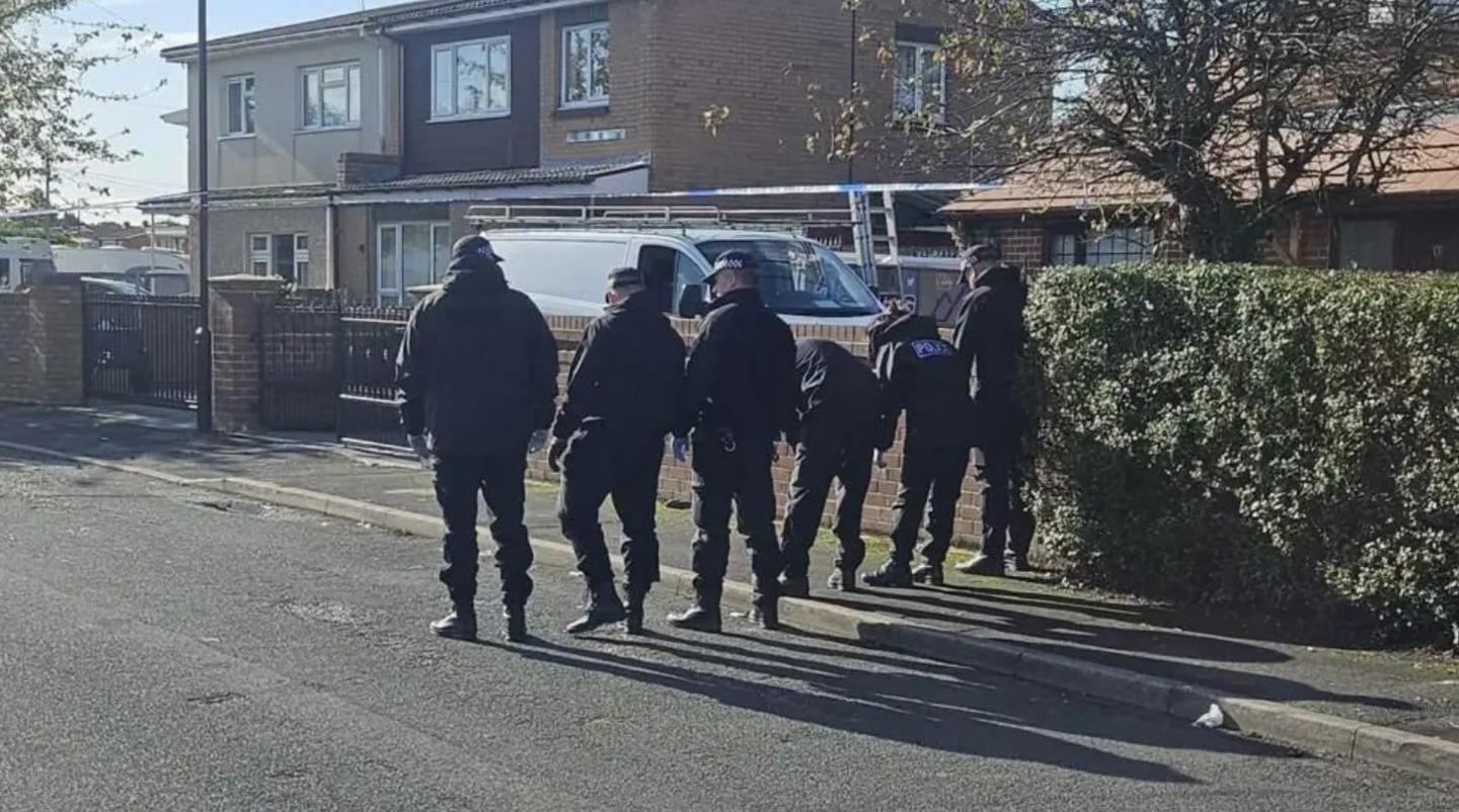 Six police officers walk down the road. Three of them are walking along the pavement, with one kneeling over to look at the ground. The other three are walking next to the group on the road. Two houses and a hedge can be seen to their right.