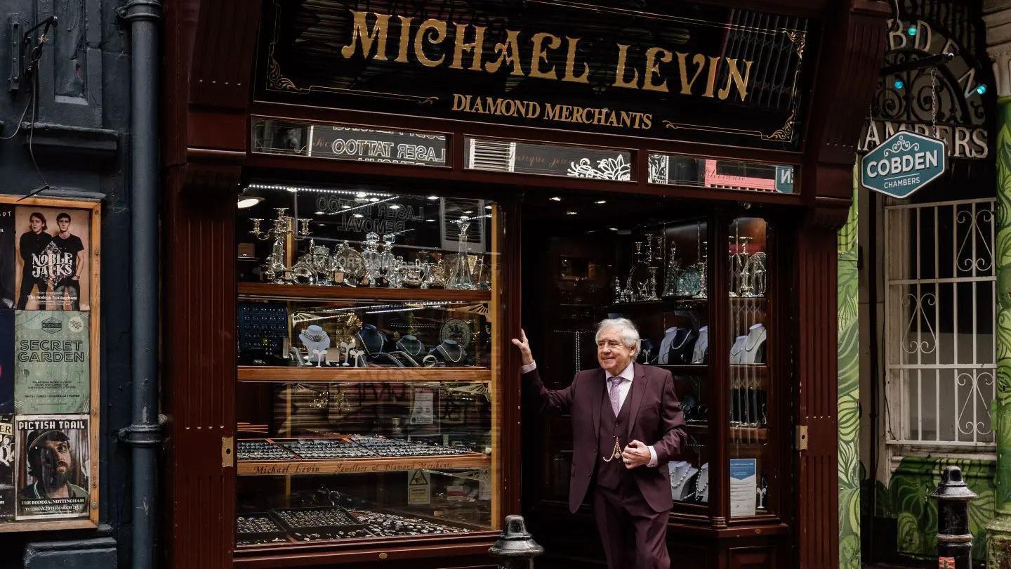 Grenville Price, in a suit, stands outside his shop, which has a traditional wood and glass frontage