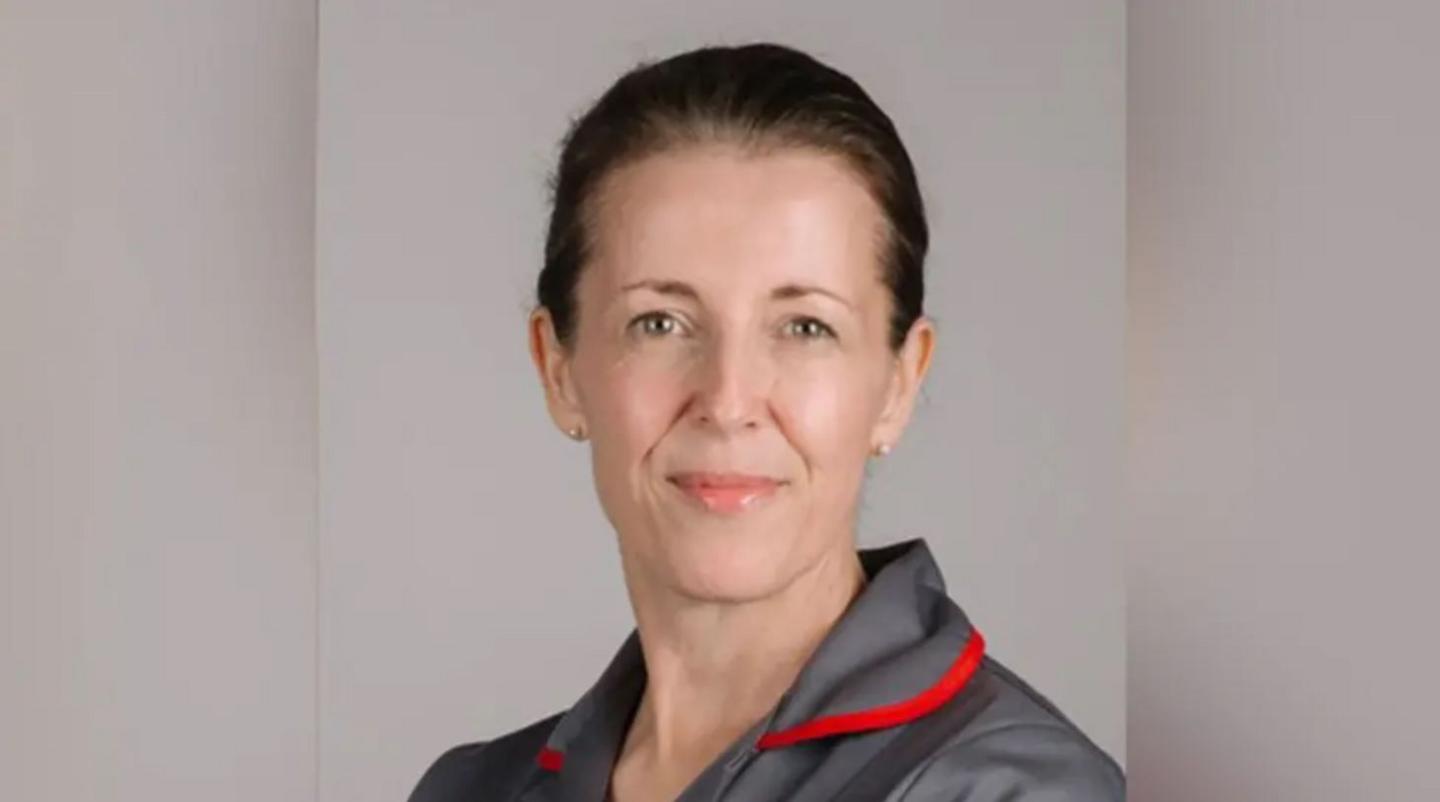 A woman with tied-back brown hair in a grey top with a red-lined collar in front of a grey background