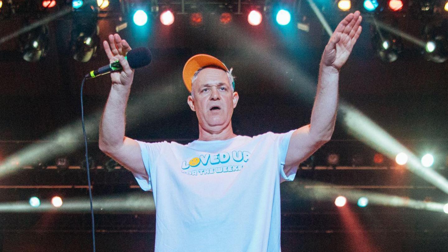 A man with grey hair in a pale blue t-shirt and orange cap with a microphone in one hand and his hands in the air, with various stage lights behind him.