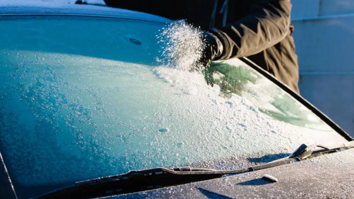 A car windscreen and bonnet covered in ice. A man in a black coat is scraping the ice off the windscreen.