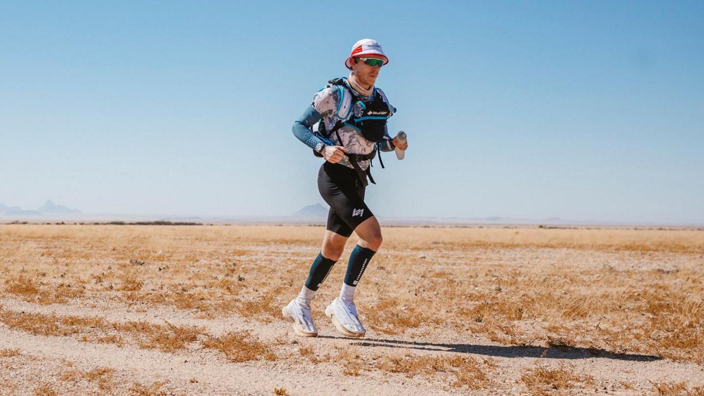 A man in running gear and a sunhat and sunglasses is running along a track in the desert. He is holding a water bottle.