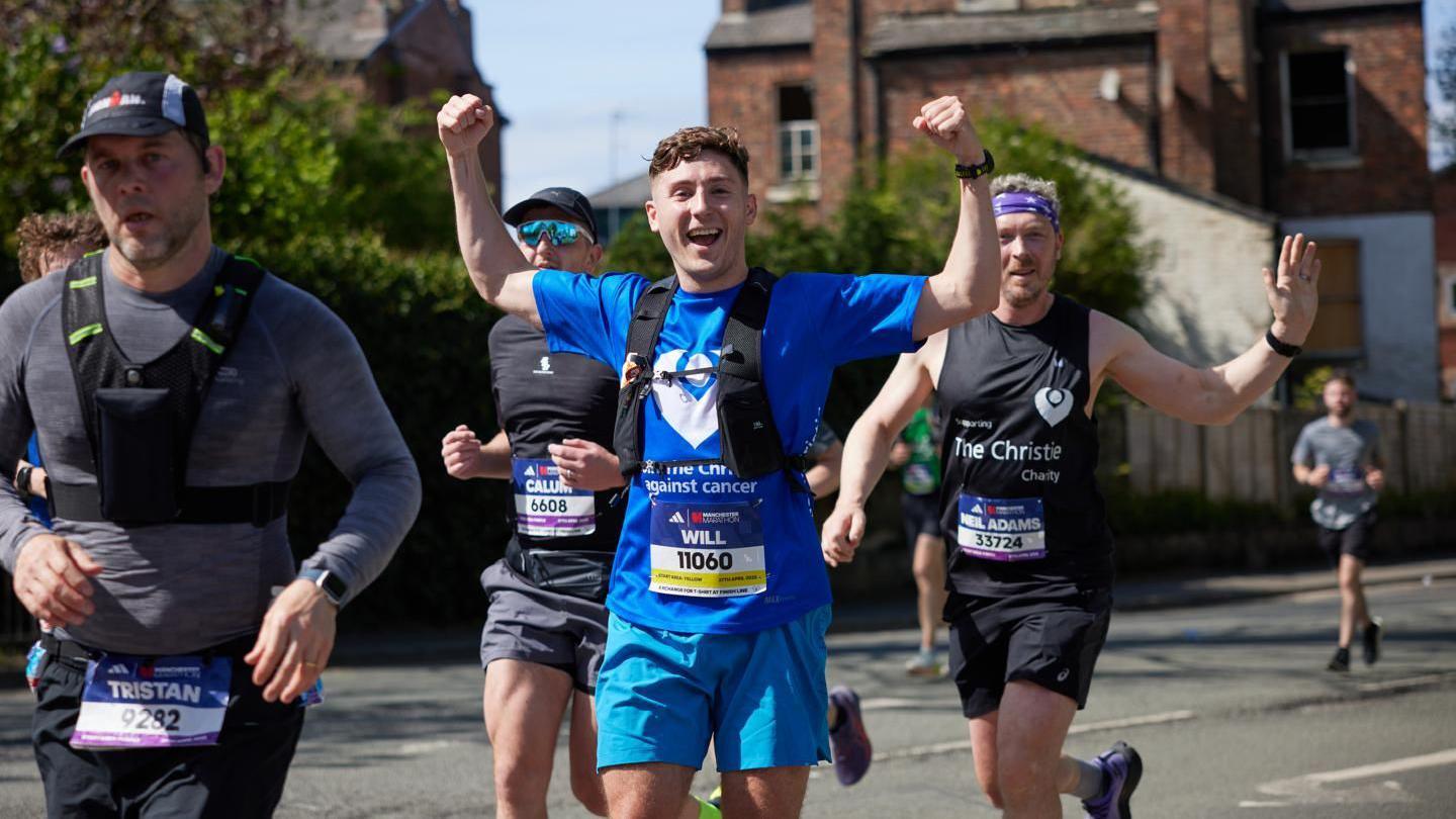 A wide shot of Will running as part of the Manchester marathon. He has his hands aloft, and is wearing a blue top and shorts.