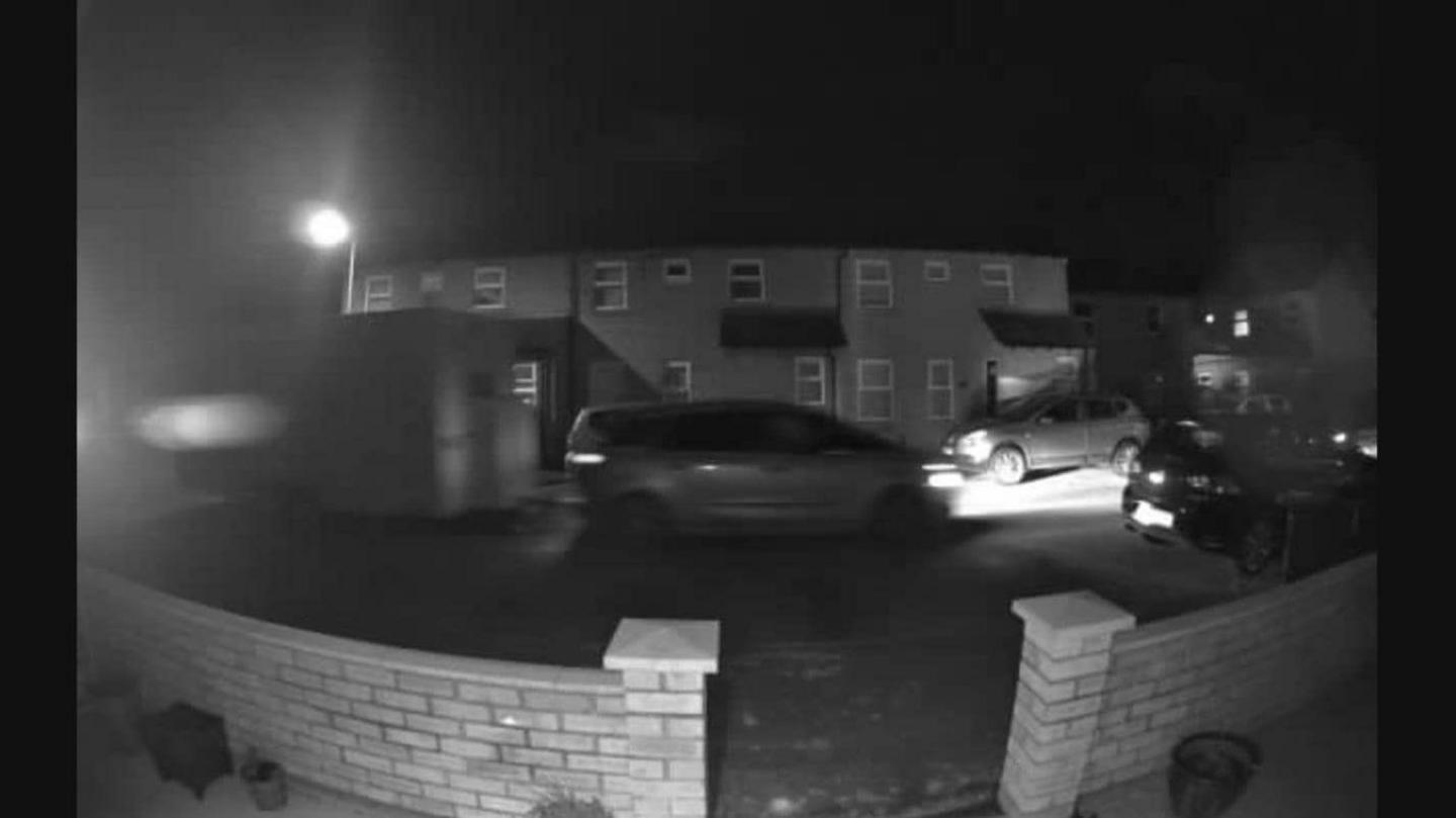 A black and white blurry still from a CCTV camera shows a car with its headlights on, towing a trailer. It is on a residential street and there are houses in the background, parked cars and a street light. In the foreground is the brick wall of the house from which the image was taken