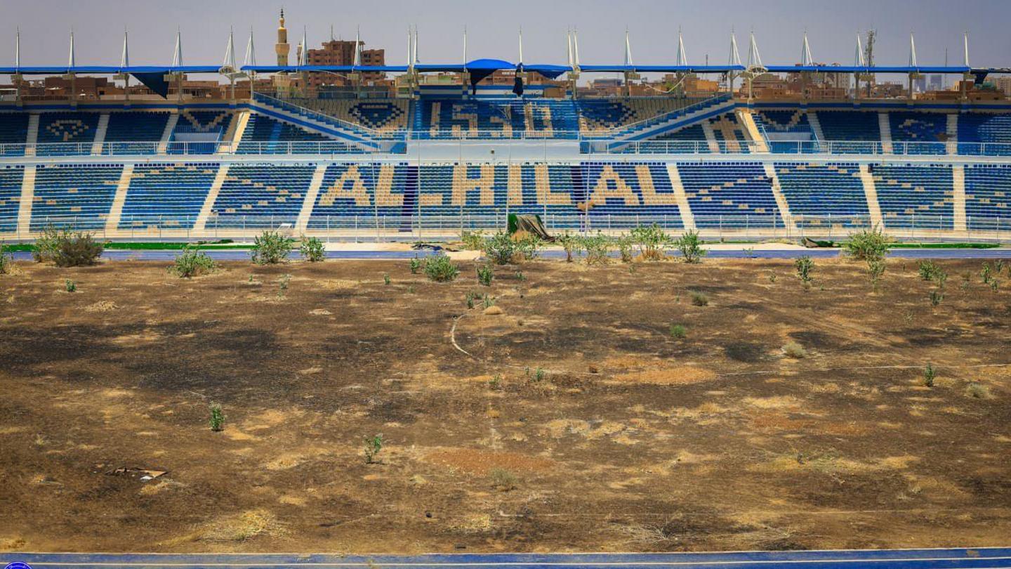 A wide view across a dirty pitch towards the stand on the opposite side. The stand and its seats are predominantly royal blue, with cream sets spelling out the name Al Hilal. There is also a blue running track visible running around the pitch and a lone minaret rising above the back of the stand. The pitch is shorn of all grass, turning it into a dry, dusty brown patch dotted with various scrubby-looking bushes on the far side of the image in front of the stand