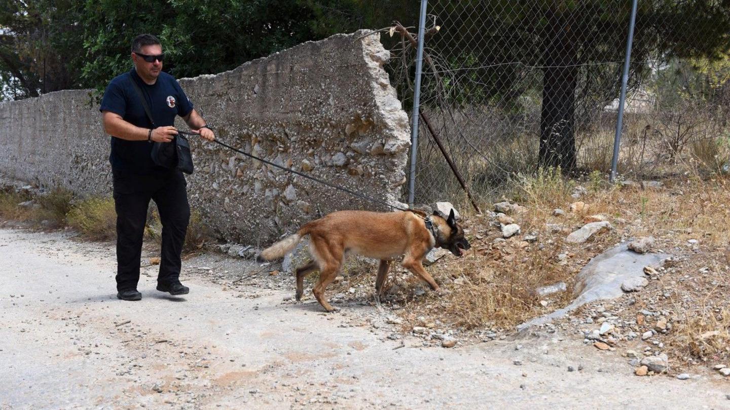 Tan coloured sniffer dog searching in an area of rubble by a fence being led by a dog handler wearing shades and dark clothing