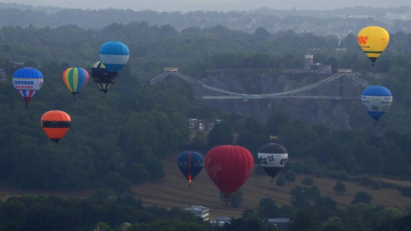 Colourful hot air balloons are flying at different heights above trees and The Downs in Bristol. There are about a dozen balloons, with blue, white, red, orange and yellow colours shining bright against the muted colours of the background. Clifton Suspension Bridge and Avon Gorge is visible in the background