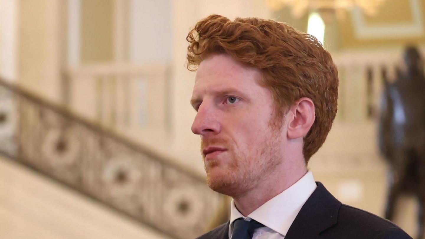 Matthew O'Toole looking sideways with the staircase of the great hall at Stormont in the background. He has red hair and wearing a black blazer and white shirt and dark green tie.