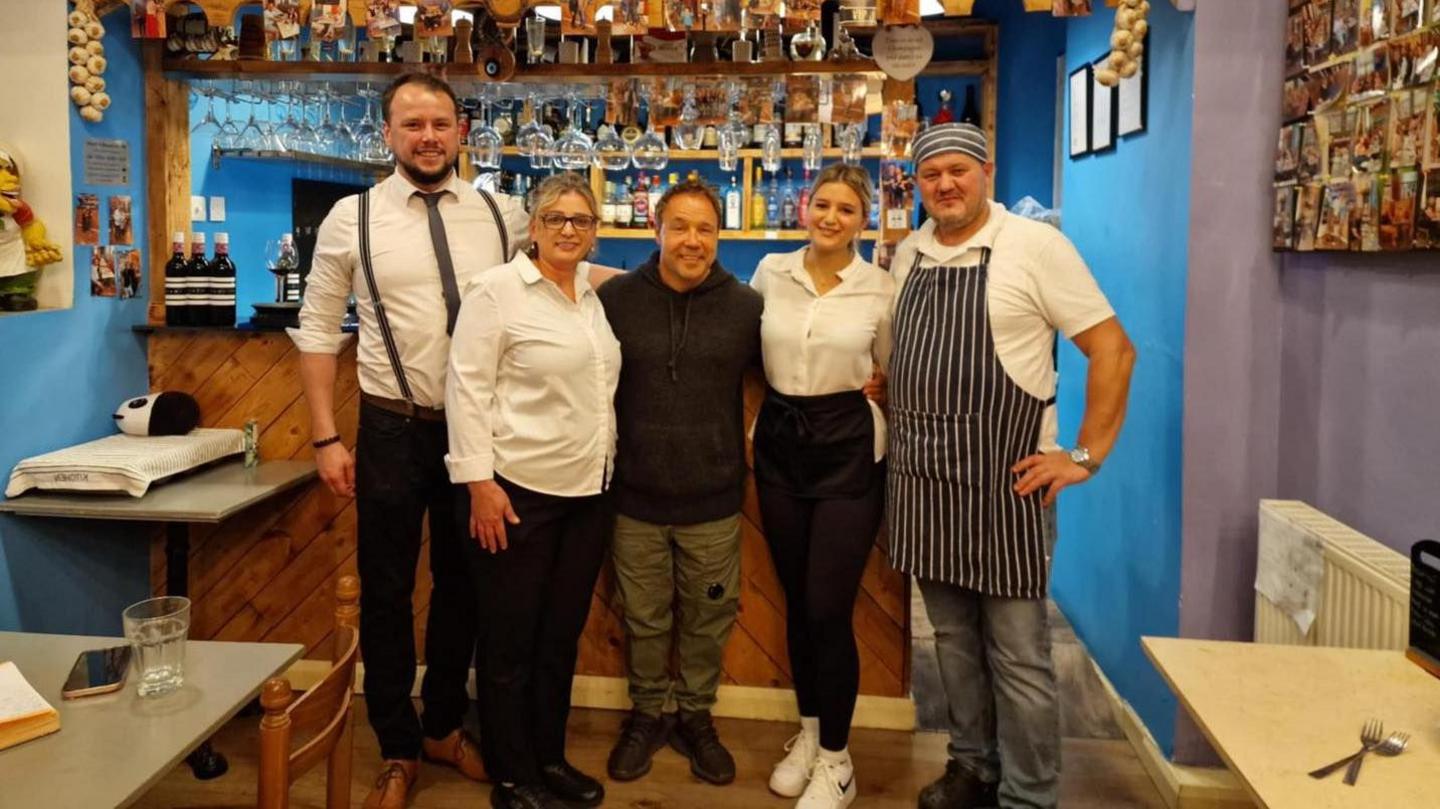 Actor Stephen Graham poses for a photograph with staff at Mr NessuNo Italian restaurant in Goole, including two men and two women. They are stood in front of a bar. The women are wearing white shirts and black trousers, while one male member of staff has a white shirt, black trousers and a grey tie and the other is wearing a blue and white striped chef's hat and apron. Stephen Graham is in the middle, smiling to the camera with his arms around both women and is wearing a black hoodie and green combat trousers.