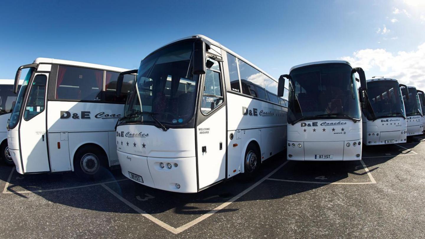 A row of white D&E coaches parked side by side in a parking area.