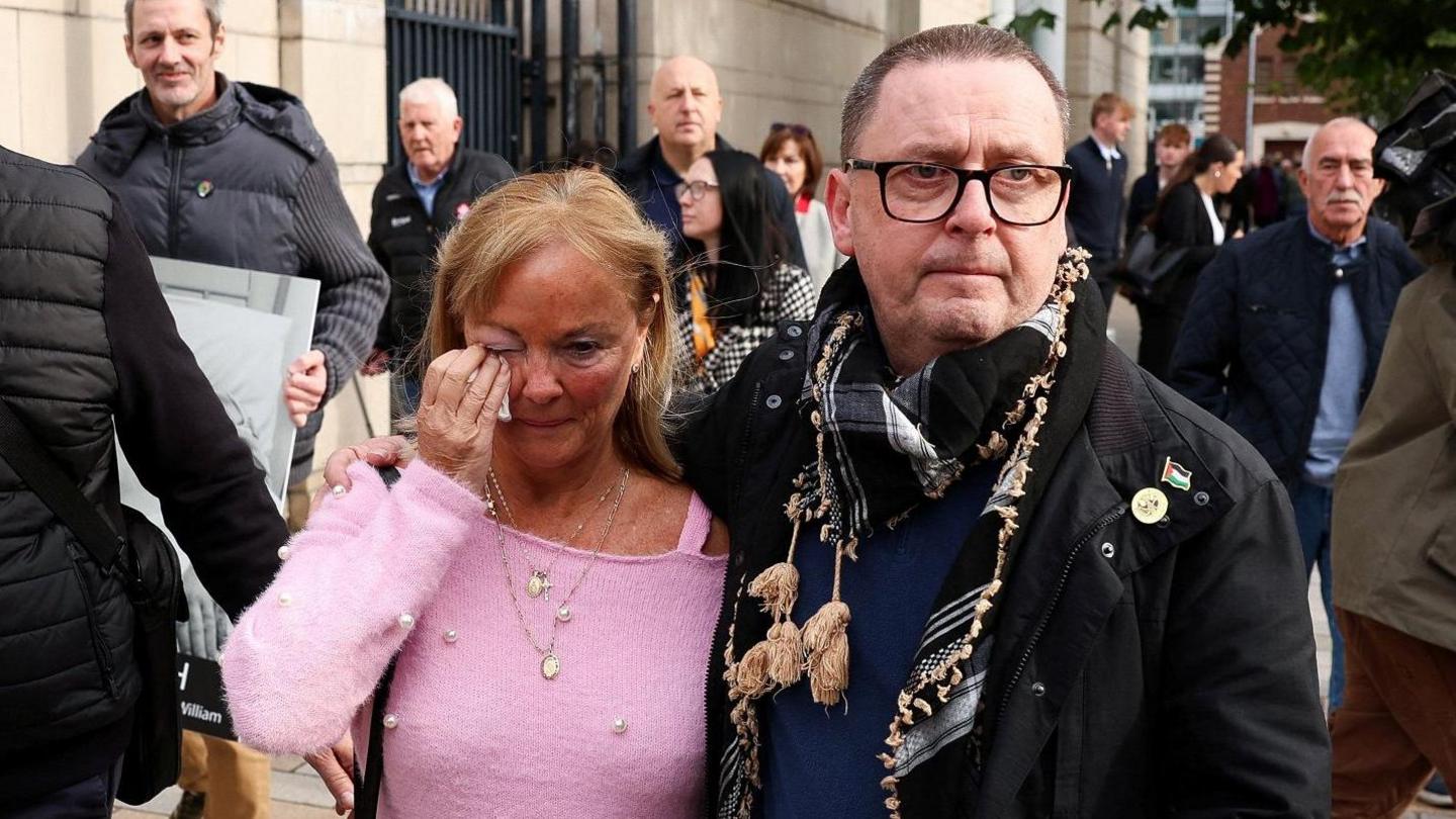 A middle-aged woman and man on a street outside a grey-bricked building. The footpath is busy with people in onlookers, with at least 10 people in the background. The woman is wearing a pink top and has blond hair. She is upset and is holding a handkerchief up to her right eye with her right hand. The man has his right arm around her shoulder, in a protective and caring way. He is wearing a black coat, has glasses and is wearing a keffiyeh around his neck.