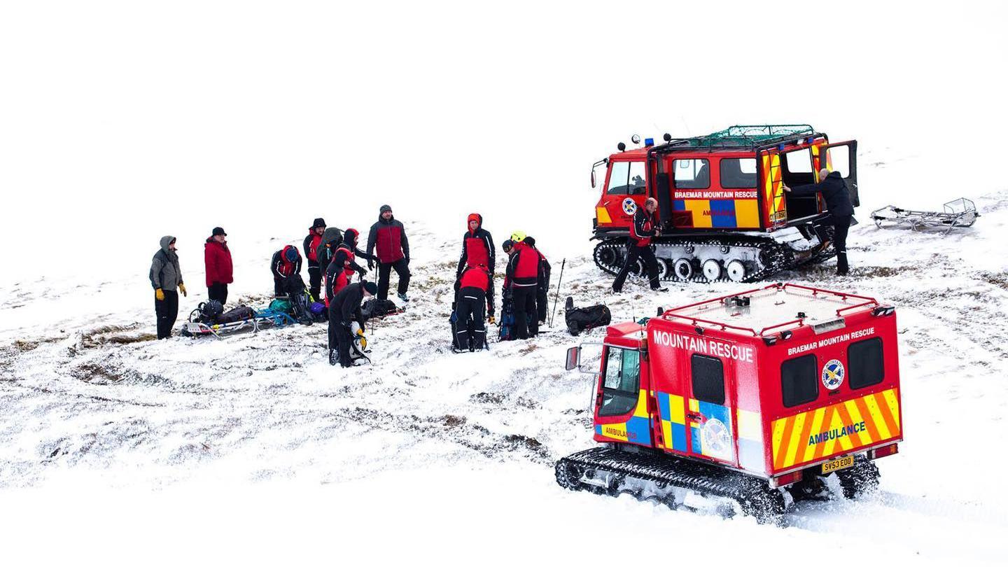 Red vehicles with tank-style caterpillar tracks, red body with white writing which says Braemar Mountain Rescue, and the word ambulance in red on blue and yellow checks, on snow, on a snowy hillside, with rescuers in bright mountain gear.