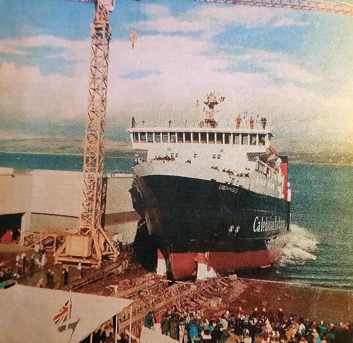 A black and white ship with red paint below the waterline on a slipway being launched. It is entering the water and crowds can be seen beside the launch ways. 