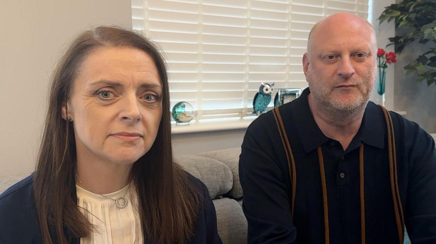Mr and Mrs Wolff sitting next to each other on a grey sofa and wearing smart clothes. They look solemn. Mrs Wolff has long dark brown hair and Mr Wolff has a shaved head and stubble.