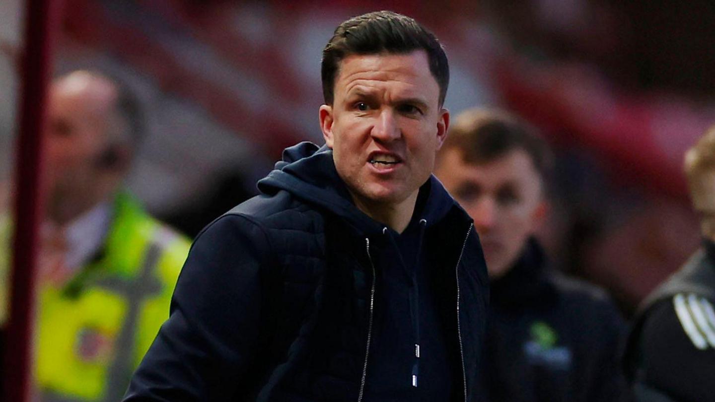 Exeter City boss Gary Caldwell, wearing a navy blue gilet over a navy blue hoodie, shouts instructions from his technical area