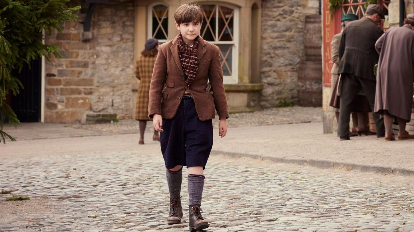 A photograph of Owen on set, wearing a brown jacket and scarf, knee-length trousers and long socks. He walks across a pebbled square, a sign that reads "Drovers Arms" can be seen on the wall in the background, partially obscured by a pine tree on the left. Three people are looking at produce on the right.