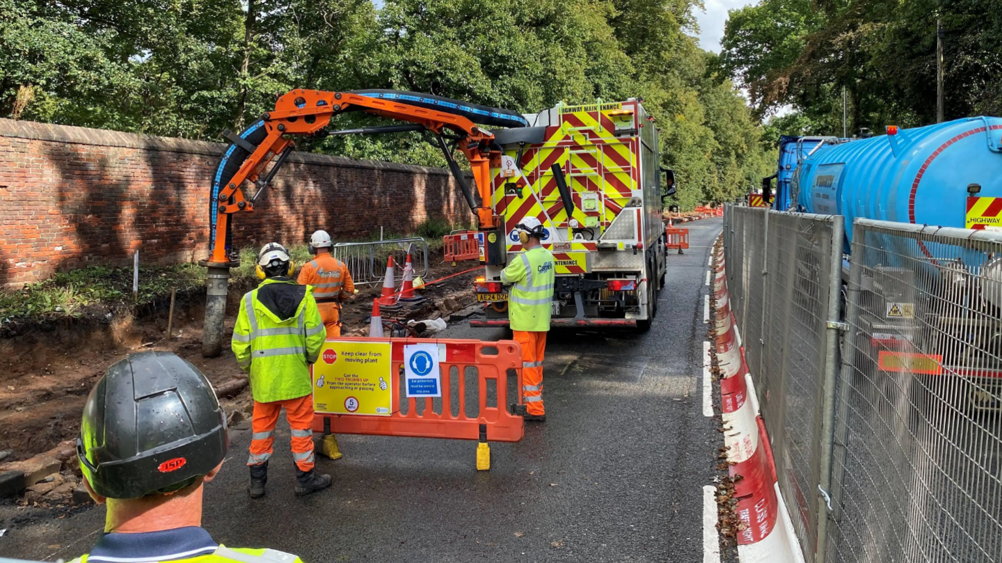 Road workers standing next to an orange barrier watching a lorry with an extended arm work on a dug out area of a road